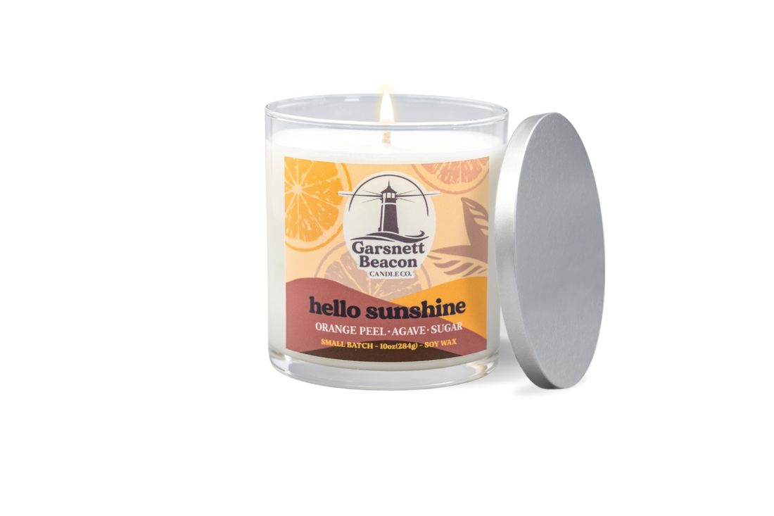 Candle labeled 'hello sunshine' on a windowsill with oranges, limes, and sunflowers.