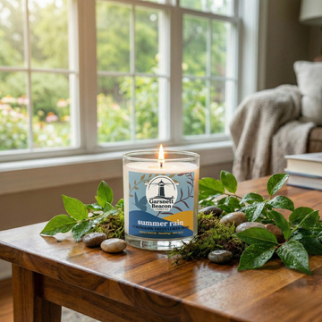 Candle labeled 'Summer Rain' on a wooden table with greenery, in a room with a window view.