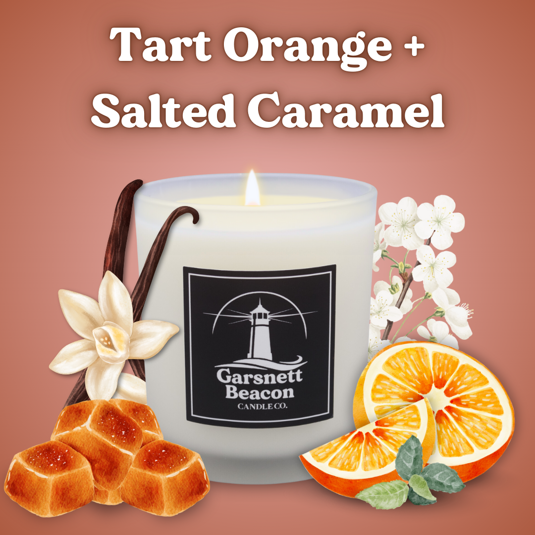 Candle with 'Garsnett Beacon' branding surrounded by vanilla bean, orange slices, and flowers on a brown background.